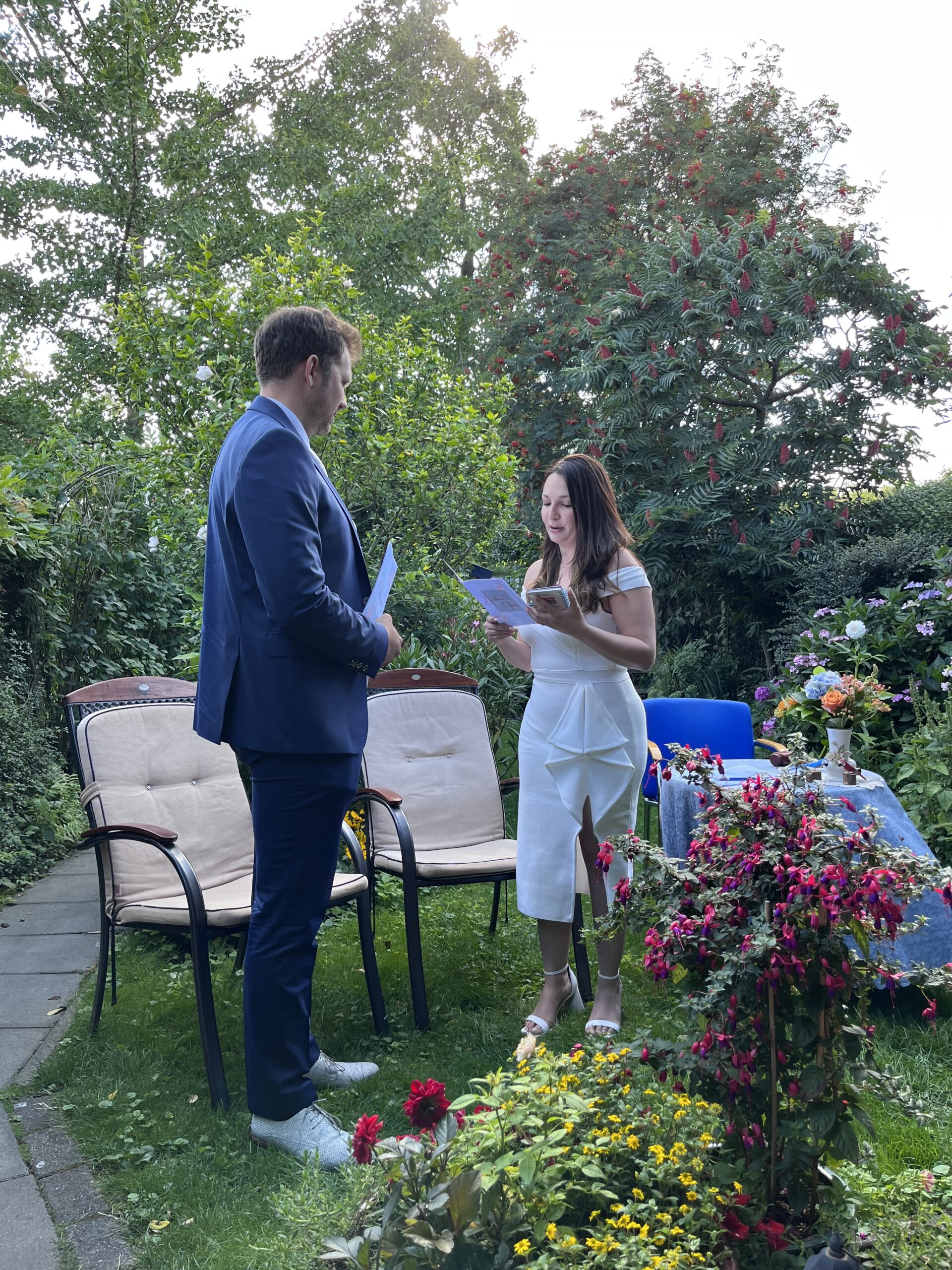 Groom and bride standing in the garden. Both of them faced each other. The bride is reading something in the paper.